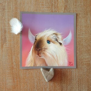 May include: A square artwork featuring a whimsical illustration of a fluffy, light brown and cream-colored creature with blue eyes and large, white ears. A white feather and a heart-shaped stone are placed near the artwork.