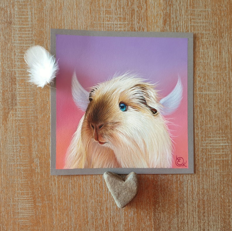 May include: A square artwork featuring a whimsical illustration of a fluffy, light brown and cream-colored creature with blue eyes and large, white ears. A white feather and a heart-shaped stone are placed near the artwork.