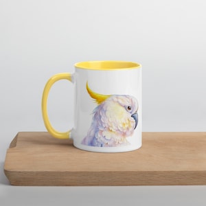 May include: A white ceramic mug with a yellow rim and handle. The mug features a watercolor illustration of a white cockatoo with a yellow crest.