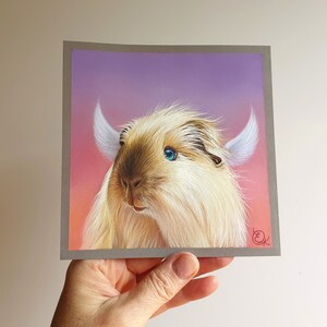 May include: A square art print featuring a whimsical creature with light brown fur, blue eyes, and white horn-like structures. The background transitions from pink to purple. The artwork is held by a hand.