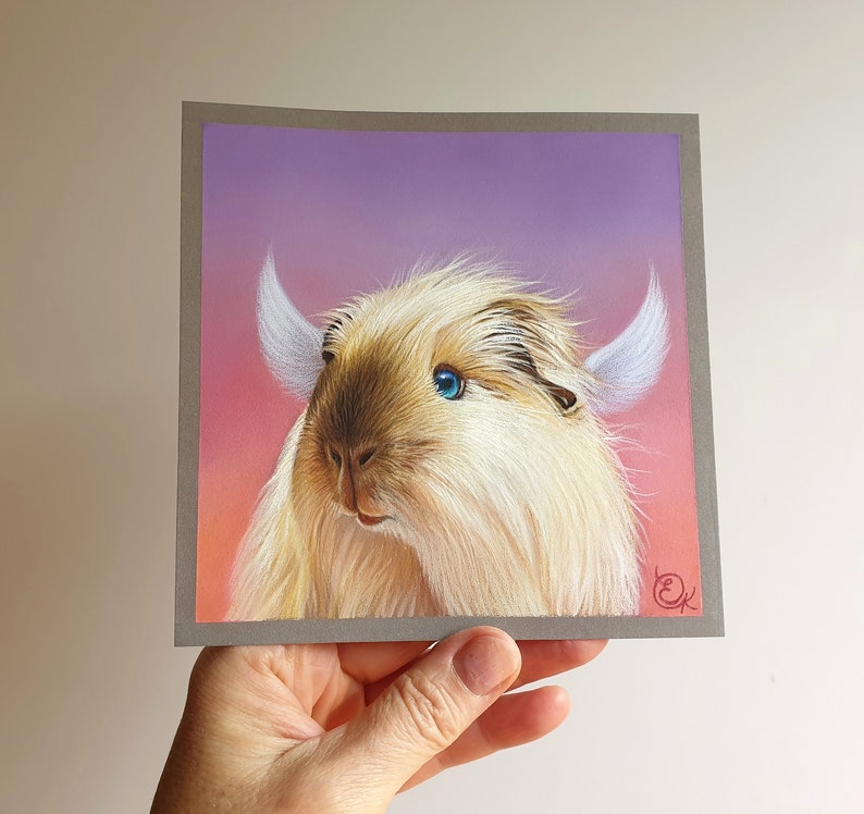 May include: A square art print featuring a whimsical creature with light brown fur, blue eyes, and white horn-like structures. The background transitions from pink to purple. The artwork is held by a hand.