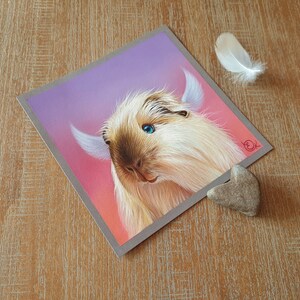 May include: A square print featuring a detailed illustration of a light brown and cream-colored guinea pig with blue eyes and large ears. The background transitions from pink to purple. A white feather and a heart-shaped stone are next to the print.