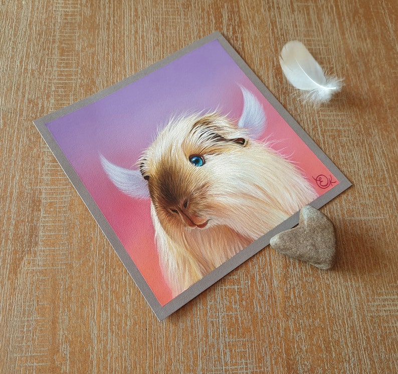 May include: A square print featuring a detailed illustration of a light brown and cream-colored guinea pig with blue eyes and large ears. The background transitions from pink to purple. A white feather and a heart-shaped stone are next to the print.