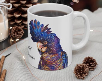Puede incluir: Taza de cerámica blanca con una ilustración de cacatúa negra. La cacatúa tiene plumas azules y moradas con detalles dorados. El texto "Galah Cockatoo" está impreso en la taza.