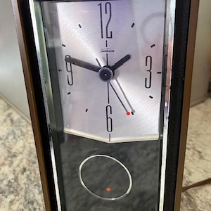 May include: Vintage Sunbeam clock with a brown frame and a silver face. The clock features black hands, a red second hand, and the Sunbeam logo. The clock has a black lower section with a circular design and a red dot.