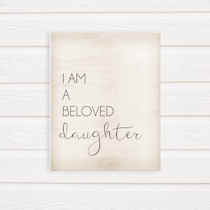 K&ouml;nnte beinhalten: Ein rechteckiges Holzschild mit dem Text "I AM A BELOVED daughter" in einer Mischung aus Serifen- und Schreibschriften. Der Hintergrund ist hellbeige, und das Schild ist vor einem wei&szlig;en Holzpaneel-Hintergrund angebracht.