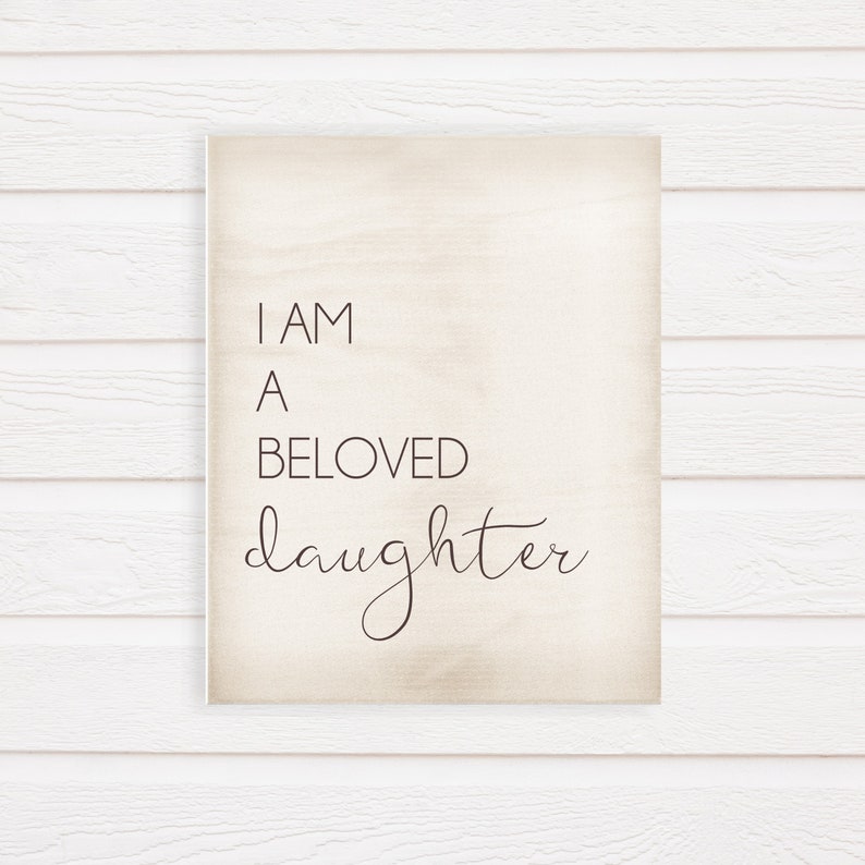 K&ouml;nnte beinhalten: Ein rechteckiges Holzschild mit dem Text "I AM A BELOVED daughter" in einer Mischung aus Serifen- und Schreibschriften. Der Hintergrund ist hellbeige, und das Schild ist vor einem wei&szlig;en Holzpaneel-Hintergrund angebracht.