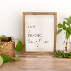 K&ouml;nnte beinhalten: Ein Holzschild mit dem Text "I AM A BELOVED daughter" in schwarzer Schrift. Das Schild steht auf einer Holzoberfl&auml;che, mit einer Holzkiste mit einer gr&uuml;nen Pflanze und einer Glasvase mit Gr&uuml;nzeug.