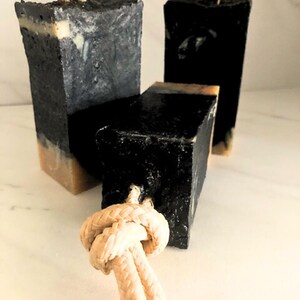 May include: Three rectangular black soaps with a light beige base and a rope knot attached. The soaps are arranged on a white surface. The beige rope knots add a decorative element to the soaps. Each soap is approximately 10 cm in height.