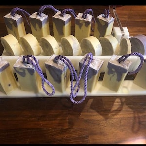 Może przedstawiać: Ręcznie robione mydła w różnych kształtach. Prostokątne mydła z fioletowymi i białymi wirami są przymocowane do fioletowych pętli z liny. Inne mydła są okrągłe lub prostokątne i jasnożółte. Mydła są wystawione na białej tacy.