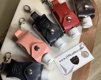 Könnte beinhalten: Fünf Lederhalter für Handdesinfektionsmittel mit silberfarbenen Metalldruckknöpfen und drehbaren Karabinerverschlüssen. Die Halter sind in Rosa, Schwarz, Rot und Dunkelbraun erhältlich, jeweils mit einer weißen Plastikflasche und einem eingeprägten Schildlogo mit dem Text I-35 Leather.