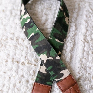 May include: A camera strap with a camouflage pattern in shades of green, black, and beige. The strap has brown leather ends and a black adjustable strap. The strap is designed to be worn around the neck.