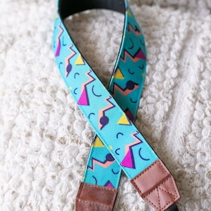 May include: A blue and pink camera strap with a geometric pattern. The strap has brown leather accents and adjustable black straps.
