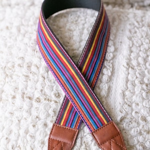May include: A colorful striped camera strap with brown leather ends and black adjustable straps.