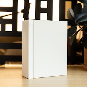 May include: A white blank book with a spine on a wooden table. A green plant is in a brown pot in the background.