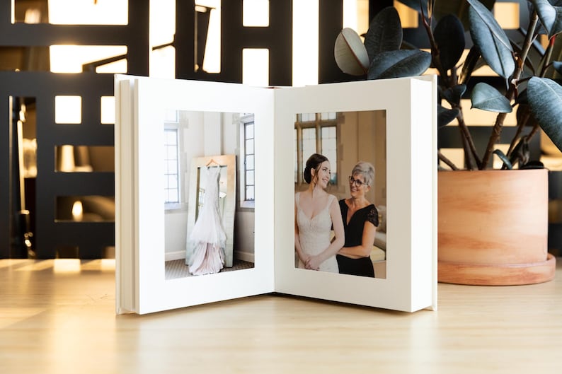 May include: A white wedding album with two photographs. The left photograph shows a white wedding dress hanging in a room with large windows. The right photograph shows a bride and her mother smiling at each other.