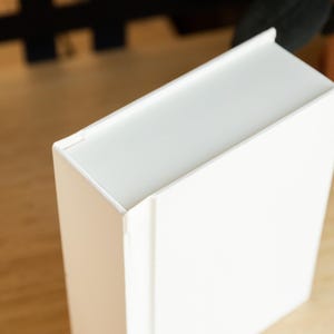 May include: A white, rectangular box with a slightly raised edge around the top. The box is sitting on a light brown wooden surface.