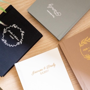 Peut inclure: Quatre livres d'or de mariage personnalisés de couleurs variées : noir, gris, blanc et beige. Chaque livre présente un texte personnalisé en écriture élégante, avec des noms et des dates, et des éléments floraux décoratifs.