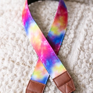Könnte beinhalten: Ein farbenfroher Kameragurt mit einem Batikmuster in Rosa, Gelb, Blau und Lila. Der Gurt hat braune Lederenden und eine schwarze, verstellbare Kordel. Der Gurt ist etwa 76 cm lang.