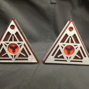 Könnte beinhalten: Zwei silberne dreieckige Metallobjekte mit einem roten Kreis in der Mitte. Der Kreis enthält ein schwarzes Symbol. Die Objekte befinden sich auf einer dunkelgrauen Oberfläche.