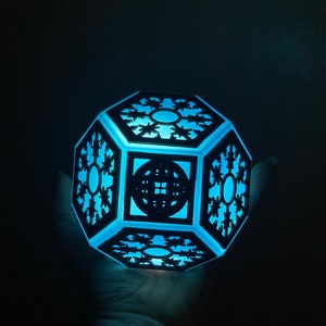 Könnte beinhalten: Eine blaue Lichtquelle leuchtet durch eine schwarze geometrische Lampe mit einem Schneeflockenmuster. Die Lampe ist aus Holz gefertigt und hat ein filigranes, kompliziertes Muster.