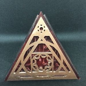 May include: A triangular wooden box with a gold-coloured design. The design features a circle with a star inside, surrounded by a geometric pattern.