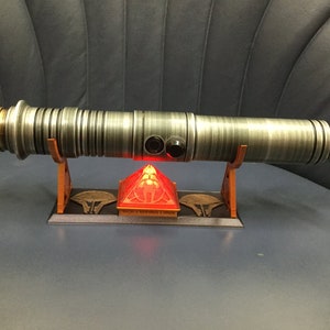 Puede incluir: Un sable de luz plateado con un acento dorado en el mango, descansando sobre un soporte de madera con una base roja iluminada.