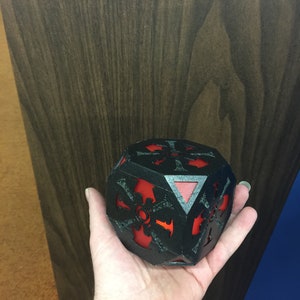 以下が含まれることがあります： 複雑なデザインが施された黒と赤の3Dプリントされたサイコロ。サイコロは人の手に握られています。