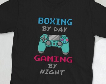 May include: Black t-shirt featuring a blue and green pixelated video game controller graphic. The text "Boxing by Day Gaming by Night" is printed on the shirt.