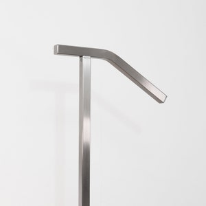 May include: A brushed metal handrail for stairs. The handrail is a simple, modern design with a vertical post and a horizontal bar that curves downward. The handrail is mounted on wooden stairs.