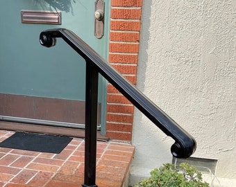 Può includere: Un corrimano in metallo nero con un design decorativo, installato su gradini di mattoni che conducono a una porta turchese. Il corrimano ha una parte superiore curva ed è sostenuto da un palo quadrato. È visibile un piccolo giardino con fiori rosa.