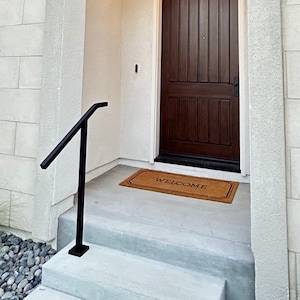 May include: A white brick house with a brown wooden door and a black handrail leading up to the front steps. A welcome mat is placed in front of the door.