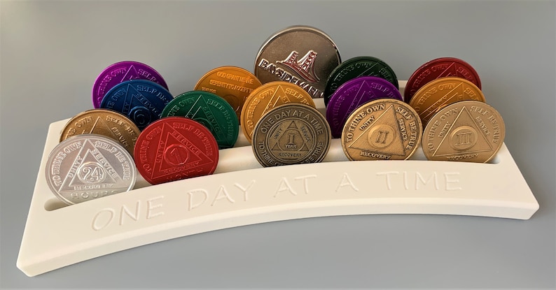 May include: A white plastic coin holder with the words "One Day At A Time" engraved on the front. The holder contains a collection of colorful sobriety coins, each with a different number of years of sobriety.