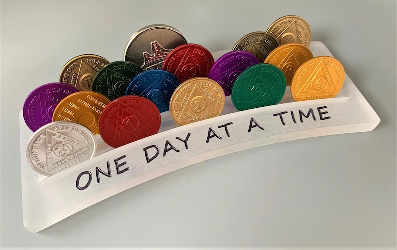 May include: A white acrylic display stand with the words "One Day At A Time" written on it. The stand holds a collection of colorful, round, metal sobriety coins with the number "1" through "4" on them.