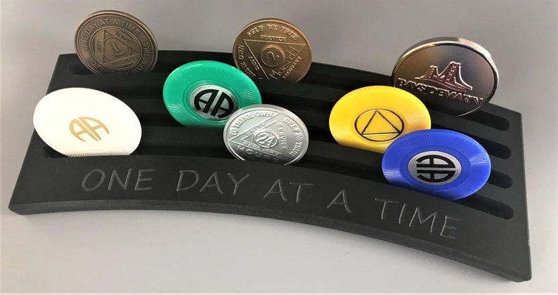 May include: A black curved display stand with the words "ONE DAY AT A TIME" engraved on the front. The stand holds seven round tokens with various designs and colors, including green, white, silver, yellow, and blue.