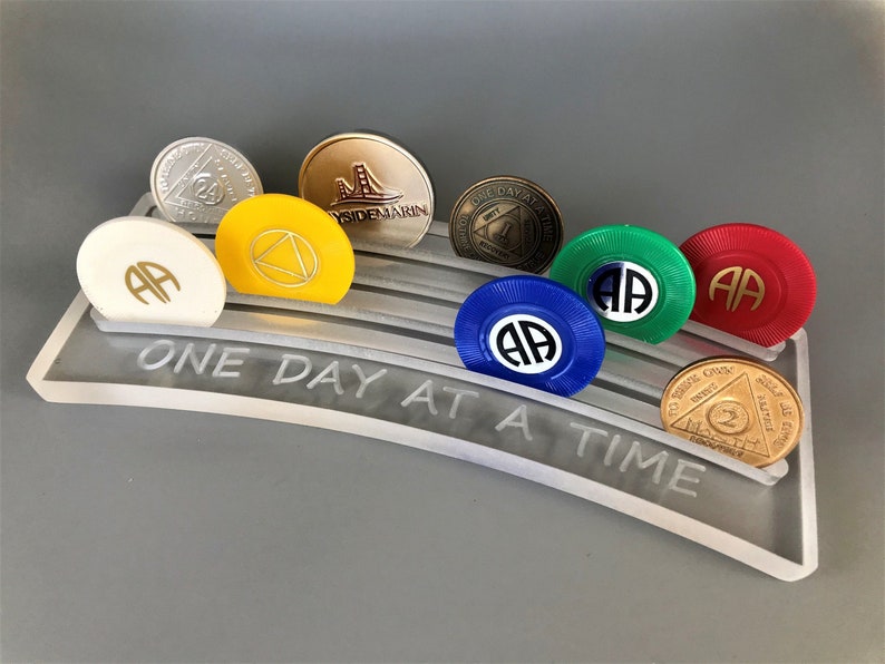 May include: A clear acrylic display stand with the inscription "One Day At A Time" holds a collection of AA sobriety coins. The coins are in various colors, including gold, silver, yellow, green, blue, and red.
