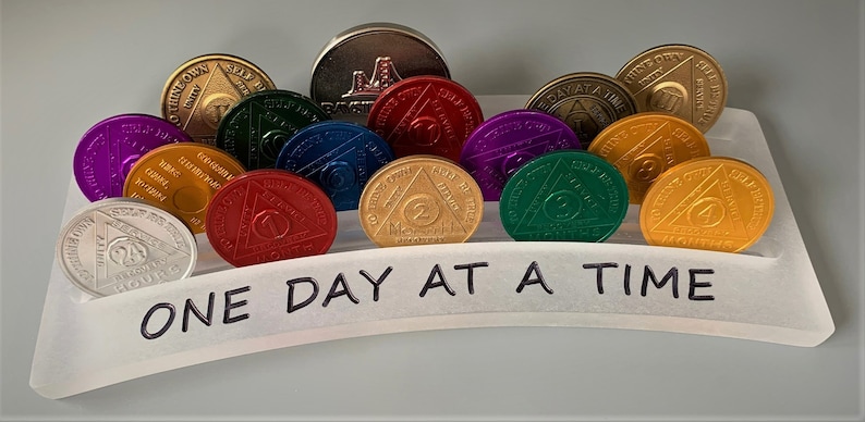 May include: A white acrylic display stand with a curved top that reads "ONE DAY AT A TIME". The stand holds a collection of gold, silver, purple, red, green, and blue round medallions with a triangle design. Each medallion has a different number on it.