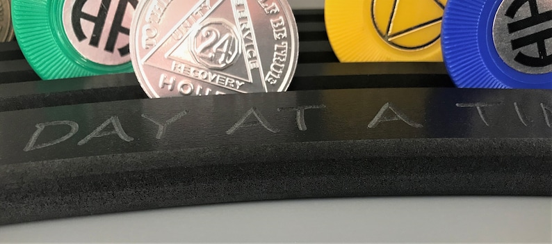 May include: A black rectangular tray with the words "DAY AT A TIME" engraved on the surface. The tray is holding several silver and gold tokens with the words "24 HOURS IN RECOVERY" and "UNITY SERVICE" on them.