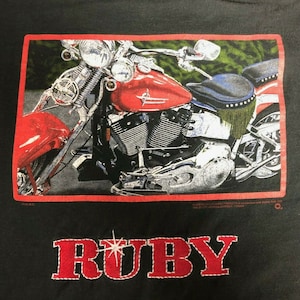 Puede incluir: Camiseta negra con un gráfico de motocicleta roja, detalles cromados y un marco rojo. La palabra "RUBY" está bordada en rojo con un diseño de estrella blanca.