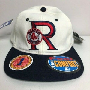 May include: A white baseball cap with a navy blue peak. The cap features a large red "R" with a Boston Red Sox logo and the text "The Comfort". The cap has a black button on top.