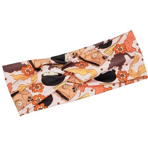 Peut inclure: Un bandeau rose avec un nœud présente un motif fantaisiste d'illustrations orange, marron et noir de chaudrons, de balais et d'étoiles.
