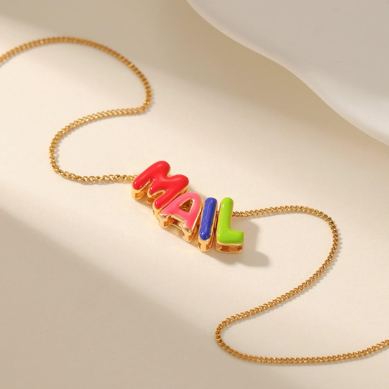 May include: A gold-colored chain necklace with the word "MAIL" spelled out in colorful, bubble-style letters. The letters are red, pink, blue, and green. The necklace is displayed on a light-colored surface.