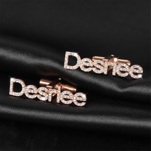 May include: A pair of rose gold cufflinks with the name "Desiree" spelled out in rhinestones.