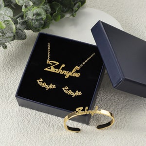 May include: A gold jewellery set in a blue box. The set includes a necklace, a pair of earrings, and a bracelet. All pieces feature the name "Zahnylee" in a cursive script.