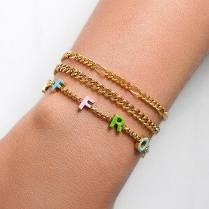 May include: A gold chain bracelet with a charm bracelet featuring the letters "F, R, O" in pastel colours.
