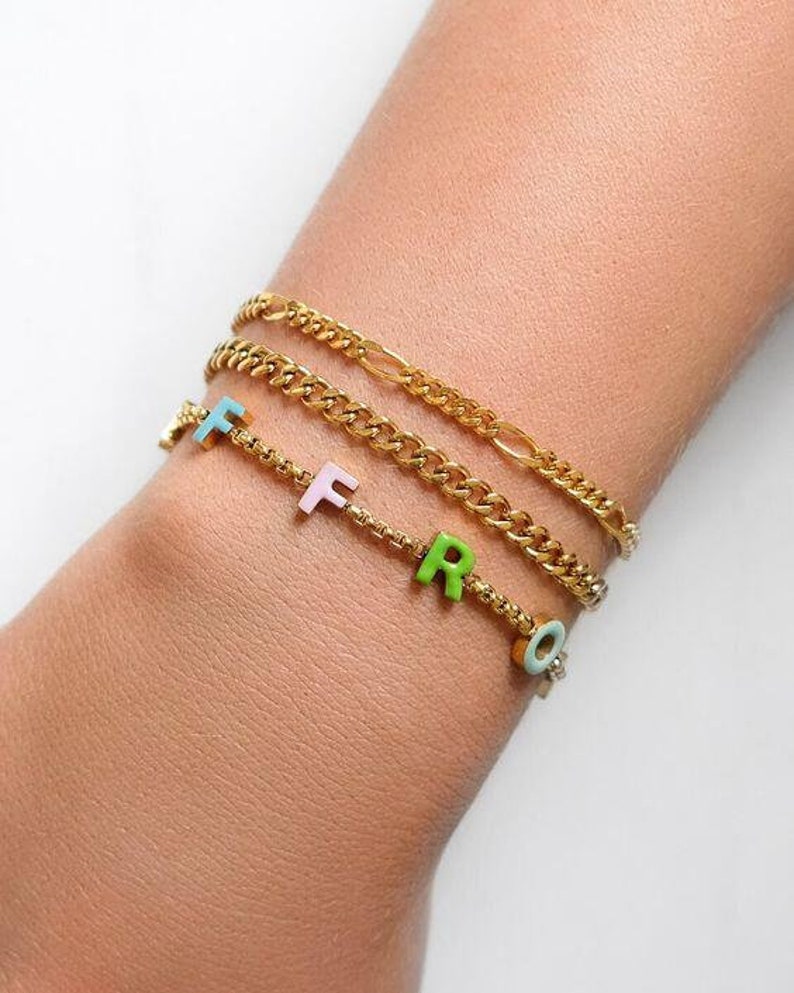 May include: A gold chain bracelet with a charm bracelet featuring the letters "F, R, O" in pastel colours.