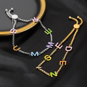 May include: Two adjustable bracelets with colourful letter charms spelling out "SUMMER" and "ZEN" on a black fabric background.