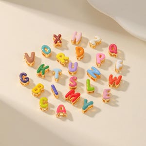 May include: A collection of colorful enamel alphabet charms in various colors, including red, blue, yellow, and green. Each letter has a gold-tone border. The charms are scattered on a light-colored surface.