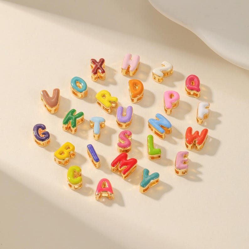 May include: A collection of colorful enamel alphabet charms in various colors, including red, blue, yellow, and green. Each letter has a gold-tone border. The charms are scattered on a light-colored surface.