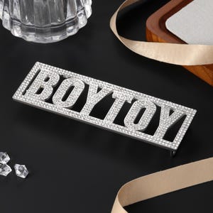 Peut inclure: Une plaque rectangulaire en métal argenté affichant le mot BOYTOY en lettres capitales. Toute la surface des lettres et le cadre environnant sont incrustés de petits strass transparents et réfléchissants qui captent la lumière.